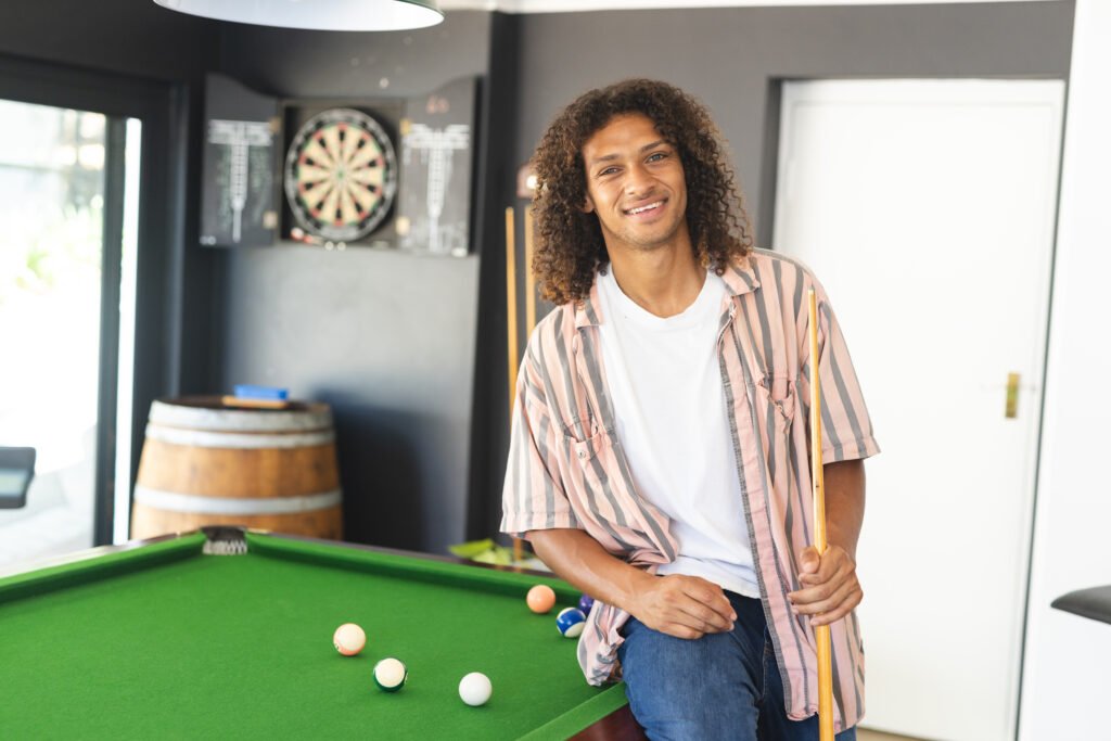 Young biracial man with curly hair stands by a pool table, smiling, with copy space. Striped shirt over a white tee, the setting suggests leisure time in a game room. Black couple arguing in bathroom wearing bath robes